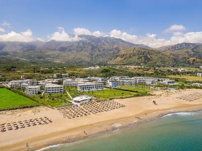Strand mit Liegen, grünen Flächen, weißen Gebäuden und Berglandschaft im Hintergrund unter blauem Himmel.