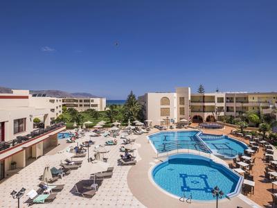 Großes Hotel mit zwei blauen Schwimmbecken, vielen Sonnenliegen und umliegenden Gebäuden, sonnig.