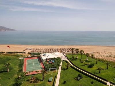 Widok na plażę z ogrodem hotelowym, kortem tenisowym i palmami pod pochmurnym niebem.
