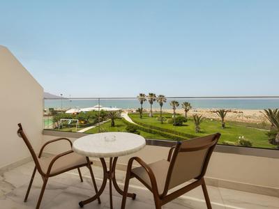 Balkon z dwoma krzesłami i stołem z widokiem na ogród i plażę pod niebieskim niebem.