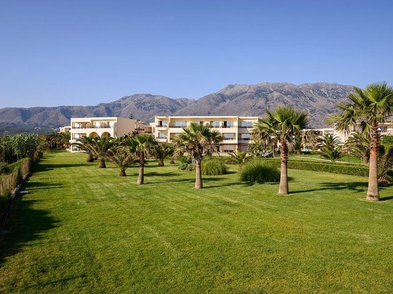 Großes grünes Gartengelände mit Palmen vor modernen Gebäuden und Bergen im Hintergrund.