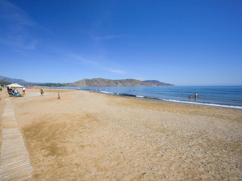 Weitläufiger Sandstrand mit blauem Himmel, ruhigem Meer und wenigen Menschen entlang der Küste.