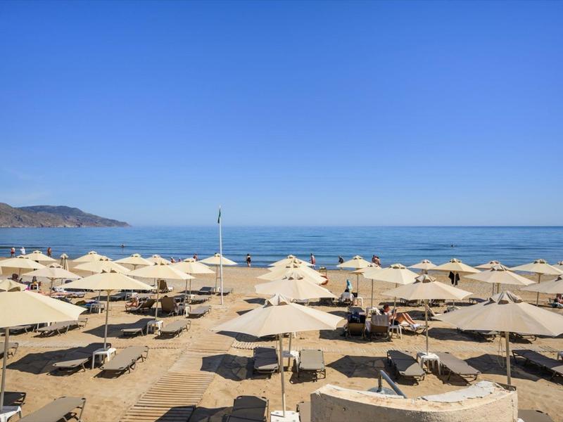 Strand mit vielen beigen Sonnenschirmen, blauem Himmel und ruhigem Meer im Hintergrund.