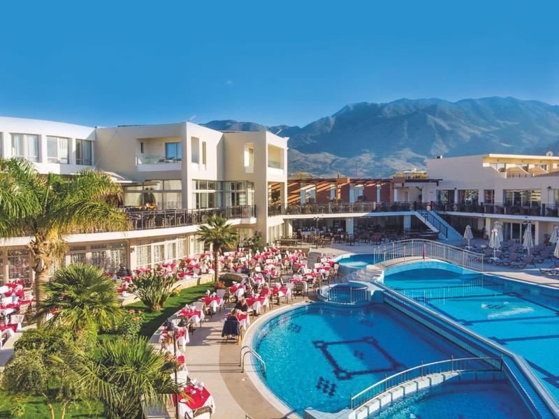 Moderner Hotelkomplex mit großem Pool, Sonnenschirmen, Palmen und Bergen im Hintergrund bei klarem Himmel.