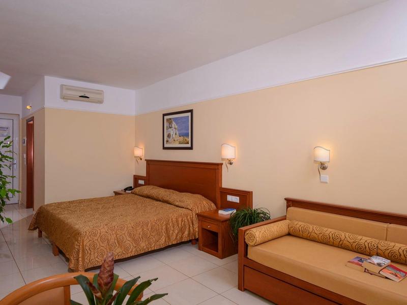 Hotelzimmer mit Doppelbett, brauner Bettdecke, Sofa, Pflanzen und schlichter Wandbeleuchtung.