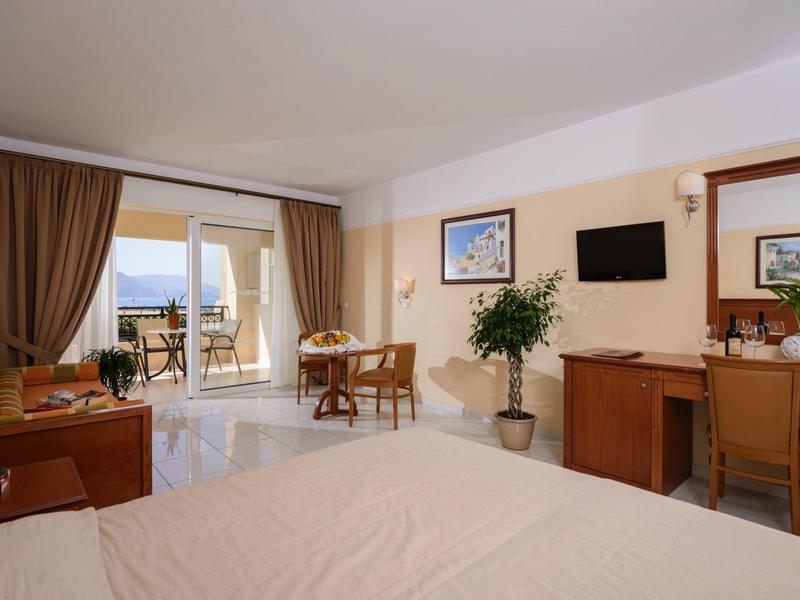 Helles Hotelzimmer mit Bett, Schreibtisch, Stühlen, TV, Pflanzen und Balkon mit Meerblick.