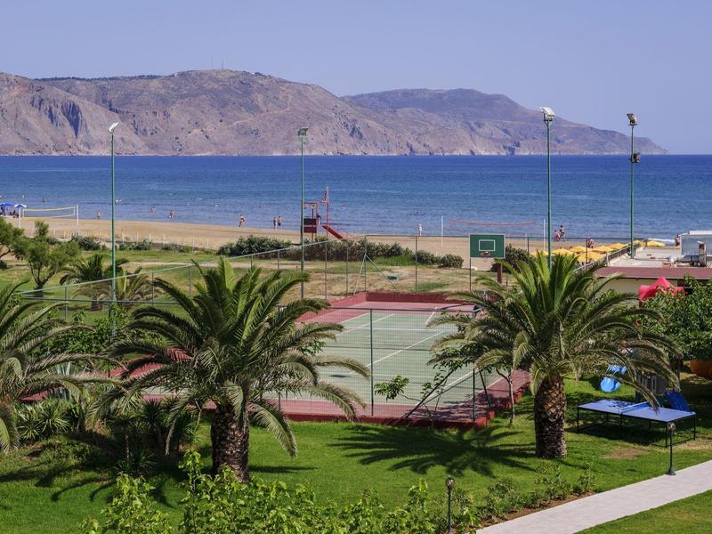 Grüne Palmen und Wiese im Vordergrund, dahinter blaue Meeresküste mit Bergen unter klarem Himmel.