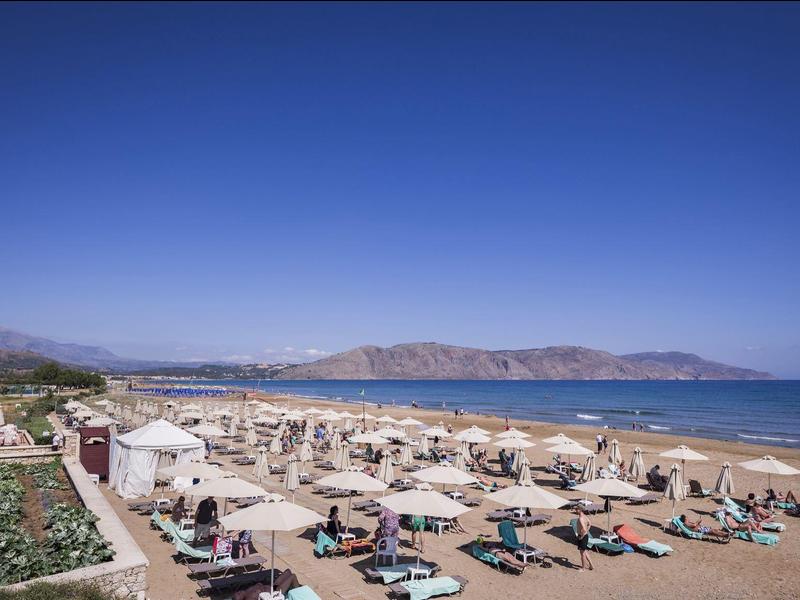 Breiter Sandstrand mit Reihen von Sonnenschirmen und Liegestühlen, blaue Sonne und Meer, Berge im Hintergrund.