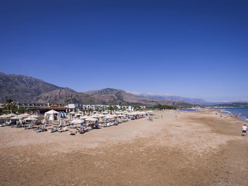 Breiter Sandstrand mit Sonnenschirmen und Liegen, dahinter Berge und klarer blauer Himmel.