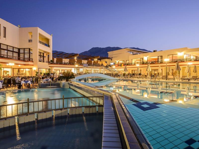Großer beleuchteter Außenpool bei Dämmerung mit angrenzenden Gebäuden und Bergen im Hintergrund.