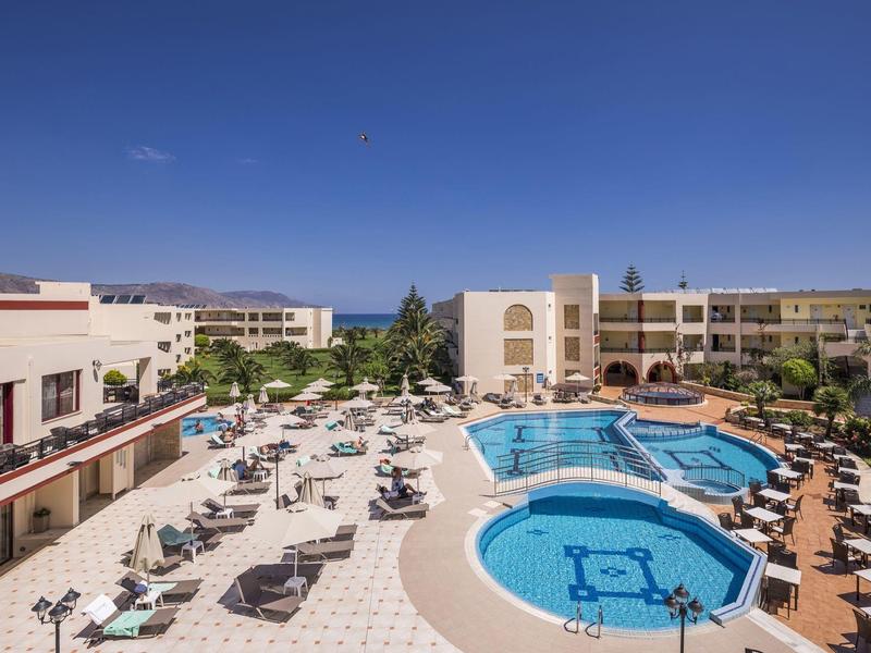 Großes Hotel mit zwei blauen Schwimmbecken, vielen Sonnenliegen und umliegenden Gebäuden, sonnig.