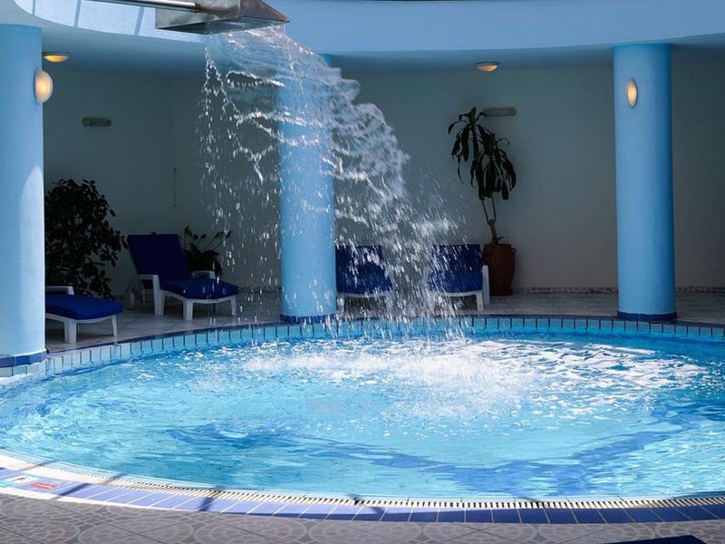 Rundlicher Hallenpool mit blauem Wasser und Wasserfontäne, umgeben von Säulen und Pflanzen.
