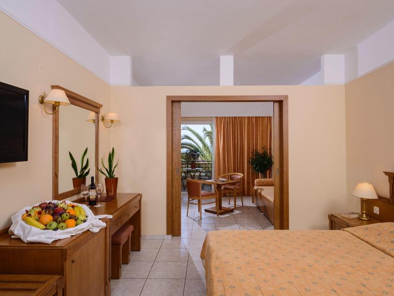 Helles Hotelzimmer mit Doppelbett, Fernseher, Holzmöbeln und Balkon mit Pflanzen und Gartenausblick.