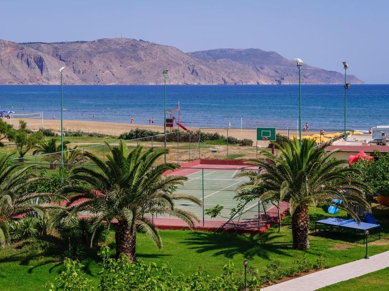 Grüner Park mit Palmen, Tennisplatz und Blick auf das blaue Meer und Berge im Hintergrund.