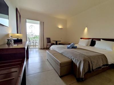 Světly hotelový pokoj s dvoulůžkem, psacím stolem, židlemi a přístupem na balkon za denního světla.