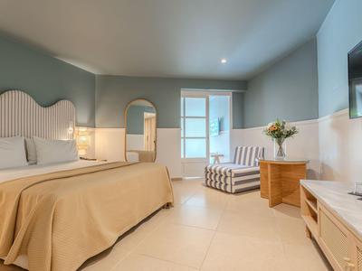 Helles Hotelzimmer mit großem Bett, gemütlichem Sessel und Fernseher an der Wand.