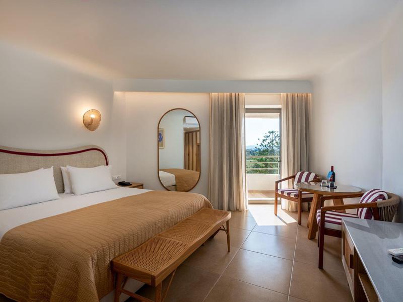 Moderní hotelový pokoj s velkou postelí, zrcadlem, balkonem a posezením s stolem a židlemi.