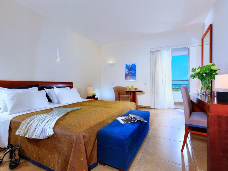 Světelný dvoulůžkový pokoj s hnědou přikrývkou, modým taburetem a výhledem na moře.