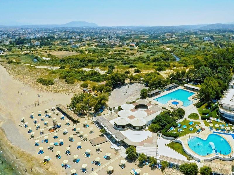 Hotelový komplex u pláže s bazény, slunečníky a výhledem na moře a hory.