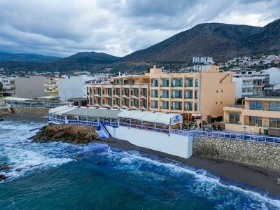 Coastal hotel by rocky shore with waves and mountainous backdrop under cloudy sky.