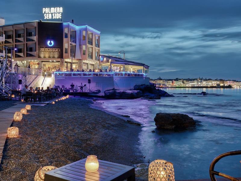 Night view of hotel by rocky sea shore with glowing lanterns on tables and pebble beach.
