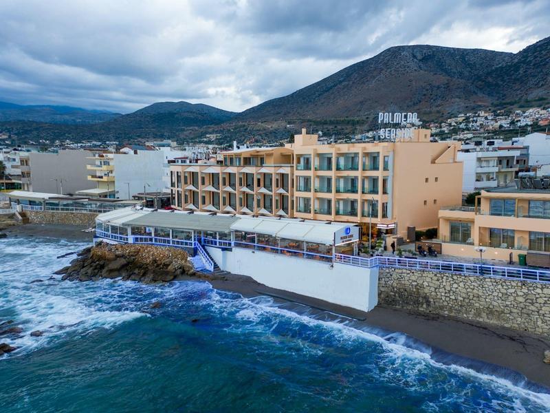Coastal hotel by rocky shore with waves and mountainous backdrop under cloudy sky.