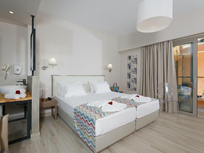 Bright hotel room with twin beds, patterned bedding, desk, and balcony access with curtains.