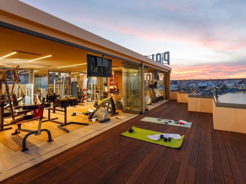 Modern gym with workout equipment on rooftop terrace overlooking city at sunset.