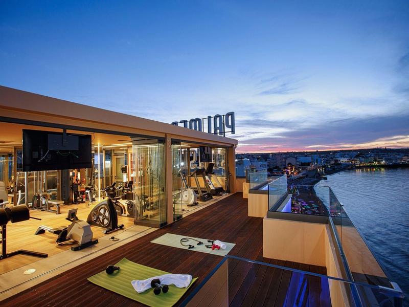 View of a hotel gym with glass walls and outdoor terrace by the water at sunset.