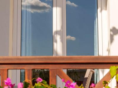 Ein offenes Fenster mit weißen Rahmen, davor grüner Strauch mit pinken Blüten und hölzerne Brüstung.