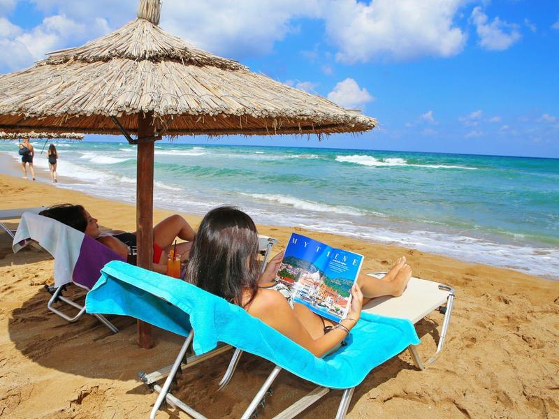Frau liegt auf blauem Strandstuhl unter Strohhut, liest Buch am sonnigen Sandstrand mit Meerblick.