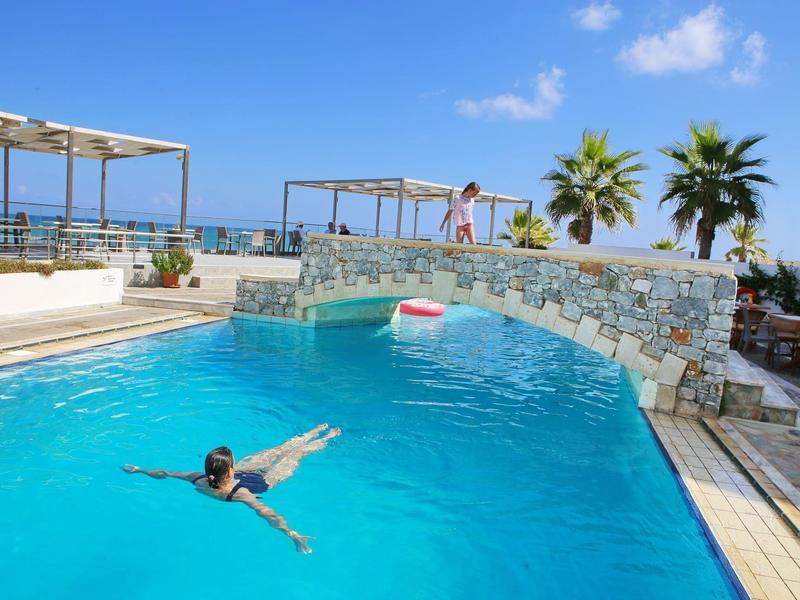 Schwimmbad mit blauem Wasser, Steinbogenbrücke, Palmen und klarem Himmel im Hintergrund.