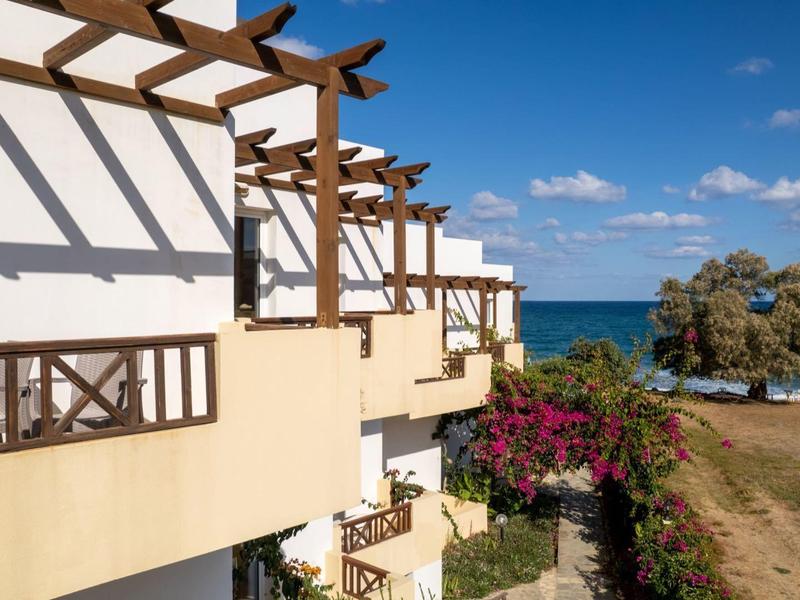 Hellgelbes Gebäude mit Holzgeländer und Pergola, blauer Himmel, Meer und Strand im Hintergrund.