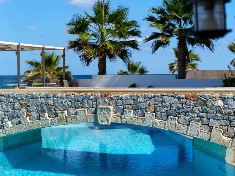 Runder Pool mit klarem Wasser, Steinmauer, Palmen und blauer Himmel im Hintergrund.