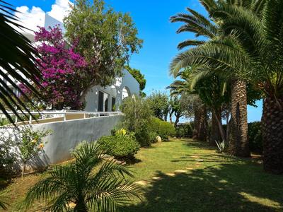 Giardino con palme e piante fiorite accanto a un edificio bianco sotto un cielo azzurro.
