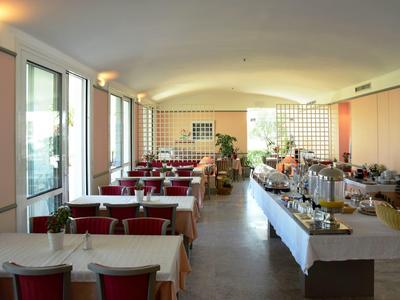 Světlá restaurace s bílými stoly, červenými židlemi a bufetovým stolem u okna.