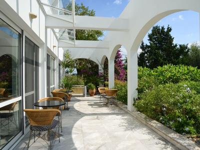 Jasně osvětlená terasa se židlemi a stoly vedle budovy a zelených rostlin.