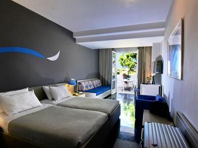 Una camera d'hotel moderna con due letti, un divano, una TV e vista su un balcone con piante.