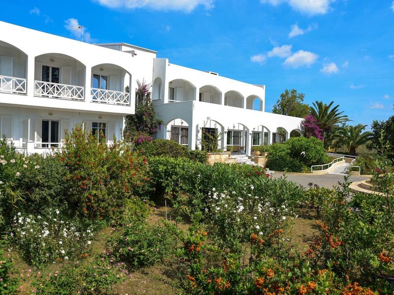 Bílá budova hotelu s balkonem a kvetoucí zahradou pod modrou oblohou.