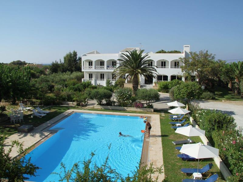 Hotel s velkým bazénem a lehátky, obklopený zelenými stromy a rostlinami.