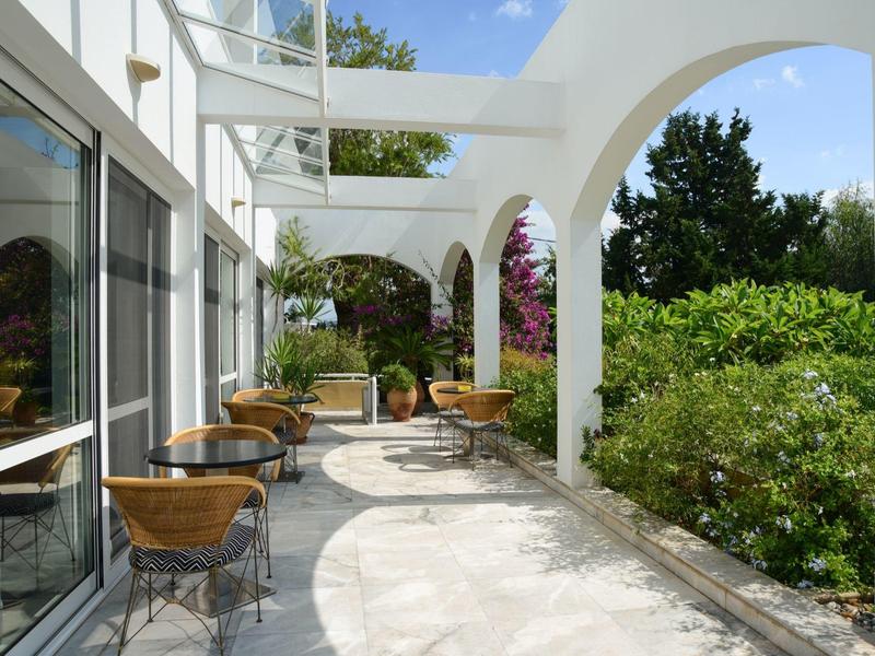 Jasně osvětlená terasa se židlemi a stoly vedle budovy a zelených rostlin.