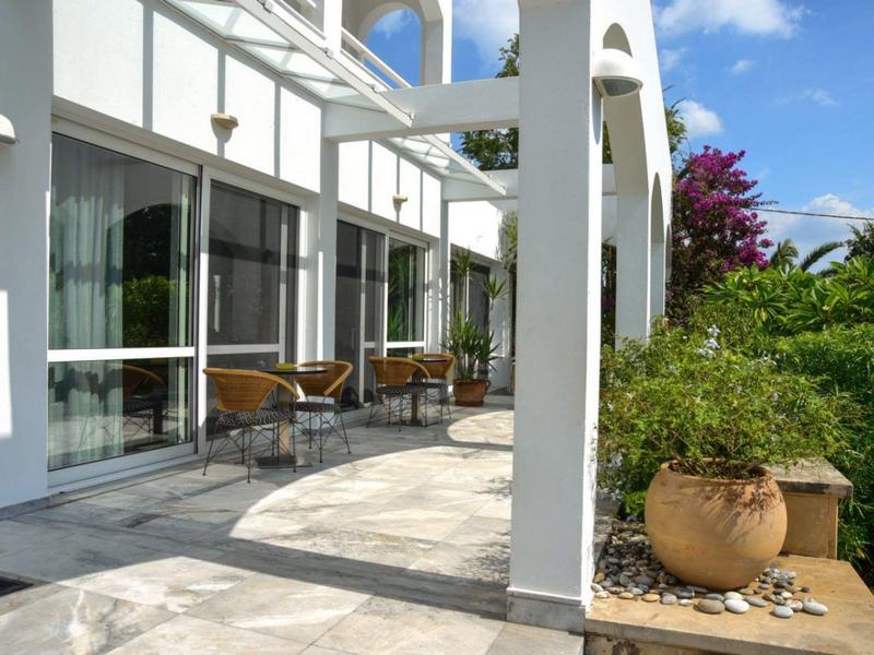 Moderní hotelový balkon s bílými sloupy, židlemi a rostlinami za slunečného dne.