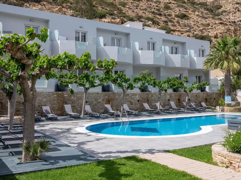 Hotel with pool, sun loungers, and two-story white buildings against a hilly backdrop.