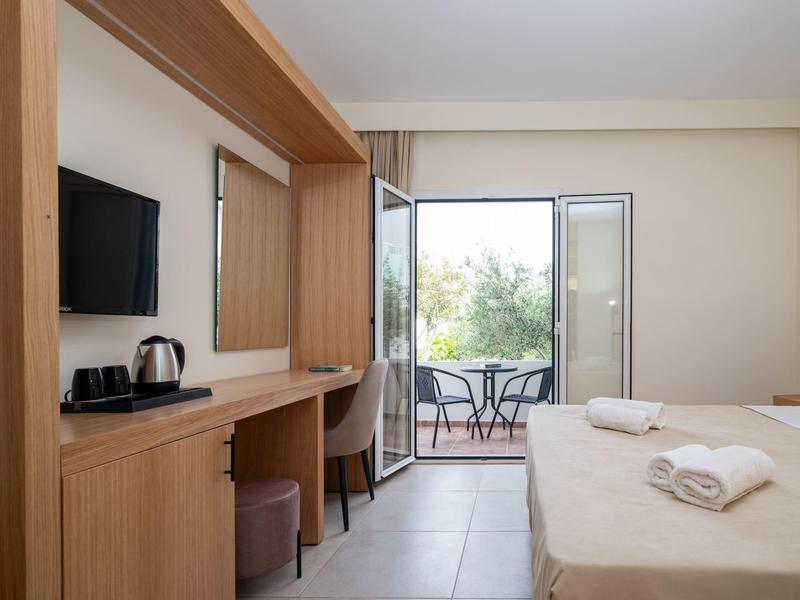 Hotel room with bed, TV, desk, and balcony overlooking greenery.