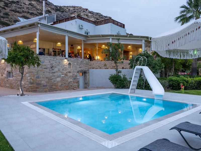 Illuminated pool with slide in front of a stone house and terrace at dusk.