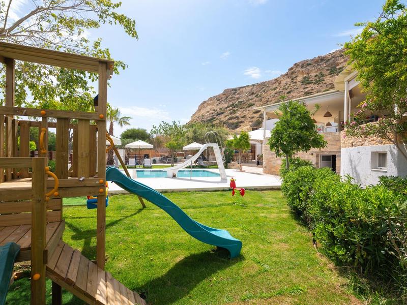 Green garden with playground and pool in front of white buildings under clear sky.