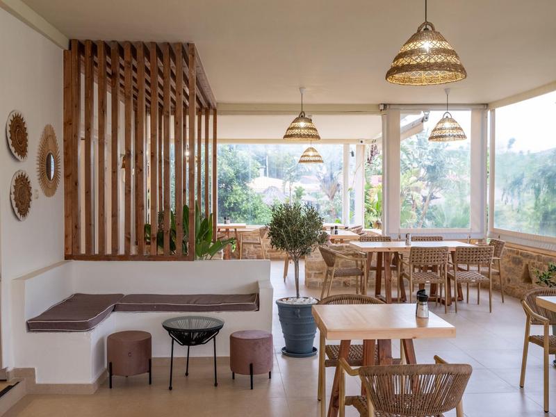 Bright restaurant with wooden furniture, plants, and large windows overlooking nature.