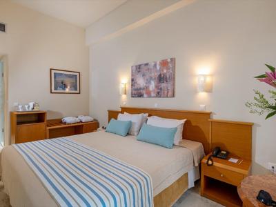 Lichte hotelkamer met groot tweepersoonsbed, blauw gestreept beddengoed en bloemendecoratie.