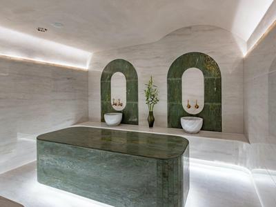 Moderne wellnessruimte met verlichte ligstoel en witte wastafels tegen groene tegelwand.