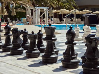 Large outdoor chess set next to a pool with palm trees and sun umbrellas in the background.